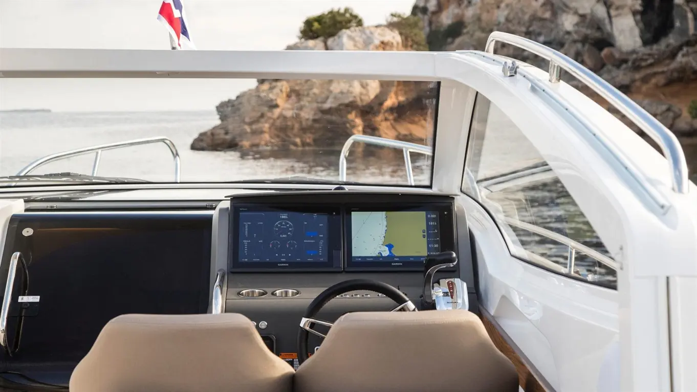 View from a boat's cockpit showing navigation screens, steering wheel, and a coastal backdrop with rocky cliffs.